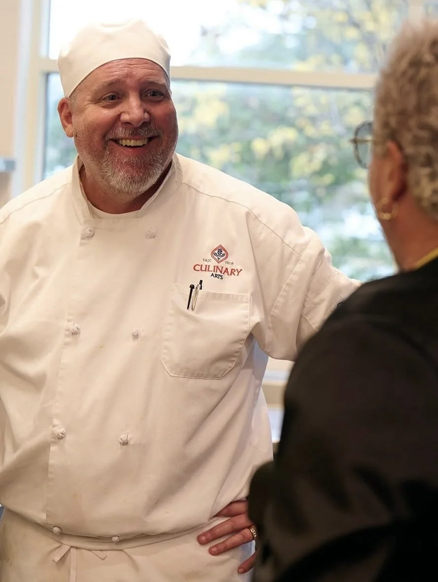 A smiling male chef wearing a white uniform and chef hat, talking with a woman, with a window showing greenery in the background.
