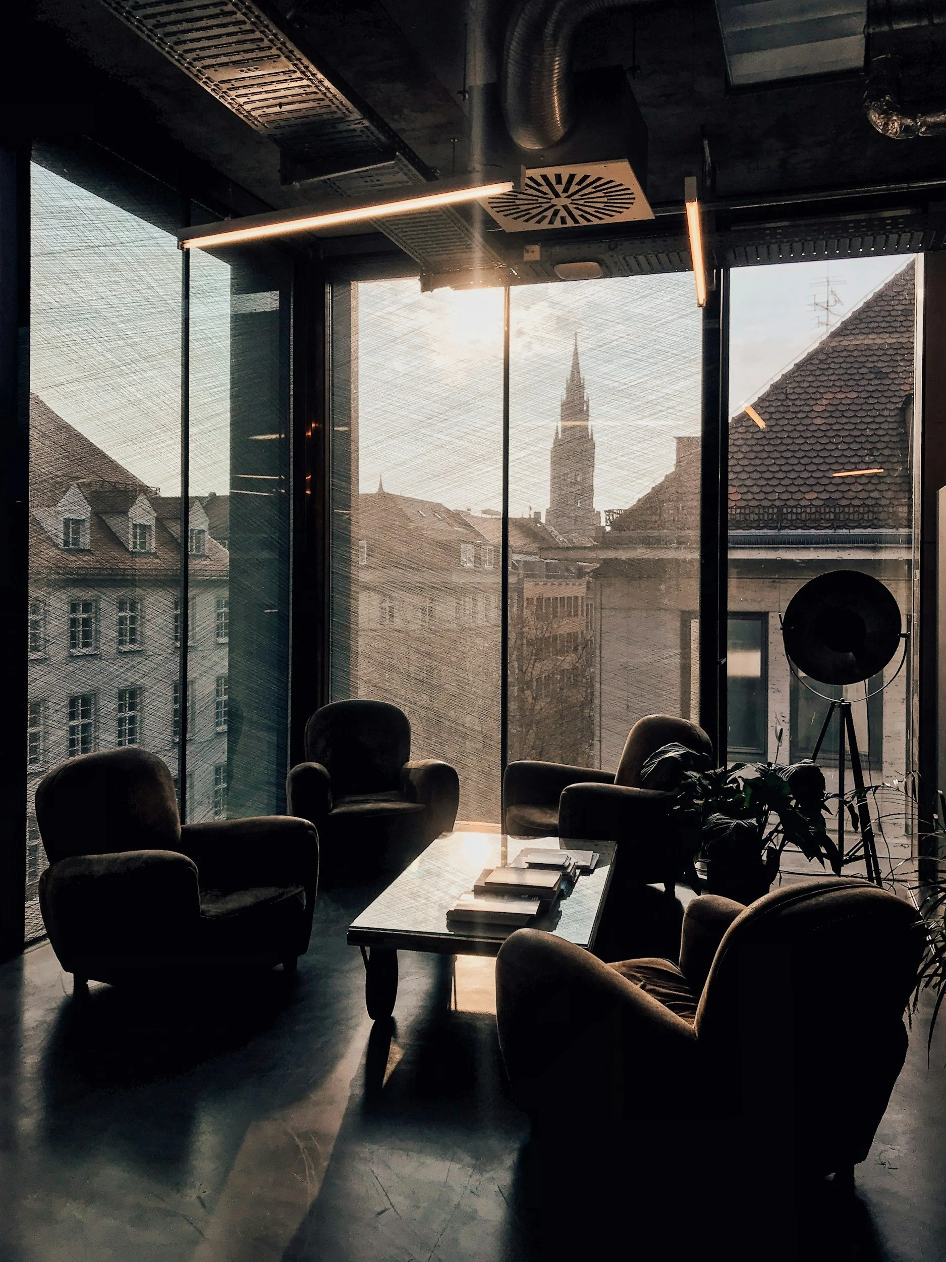 A modern interior office space with a glass wall overlooking a cityscape with traditional European-style buildings and a church steeple, with a row of armchairs, a coffee table with books, and a large floor-standing lamp.
