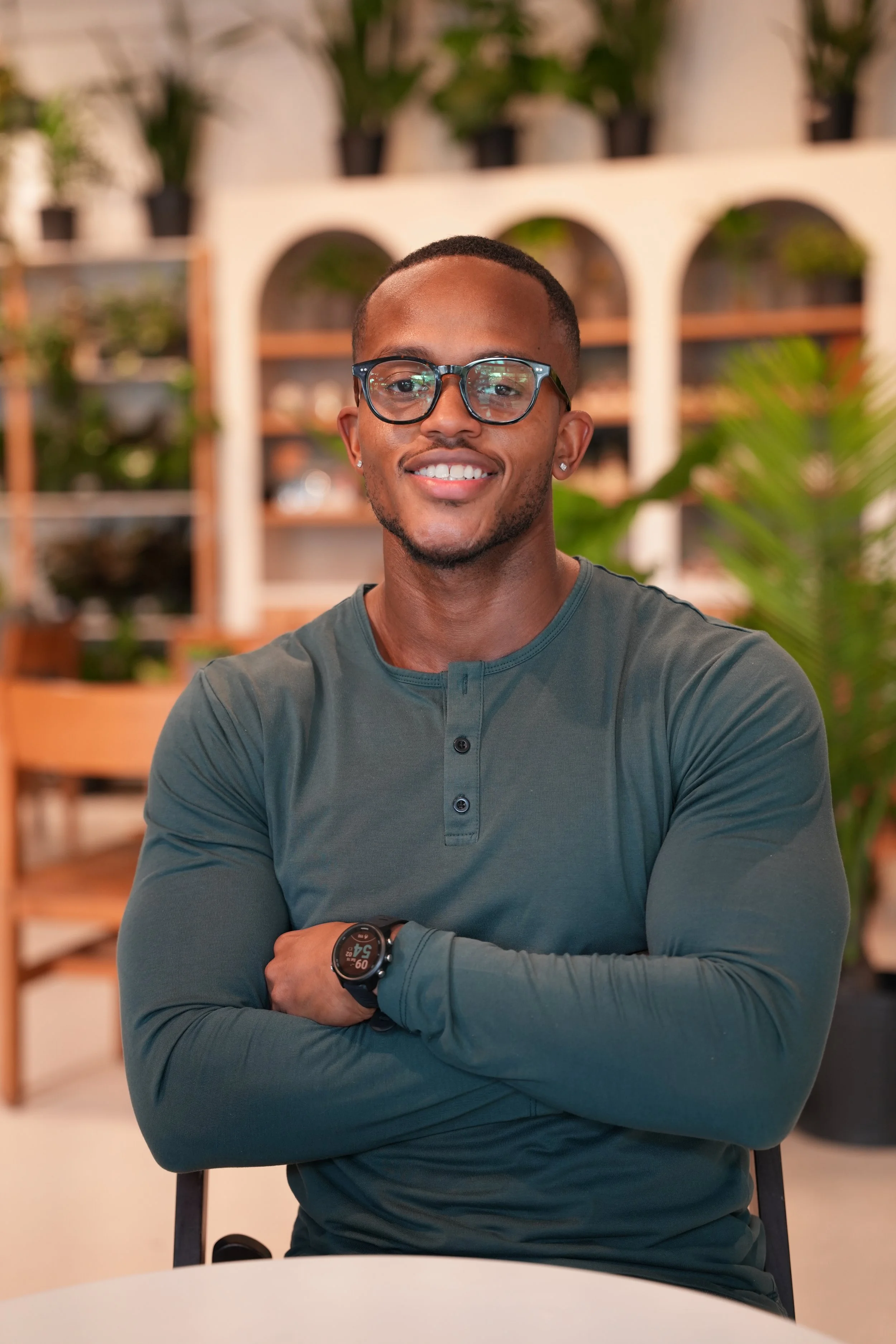 A smiling young man with glasses, a watch, and earrings, sitting with arms crossed in a modern indoor space with shelves and plants in the background.