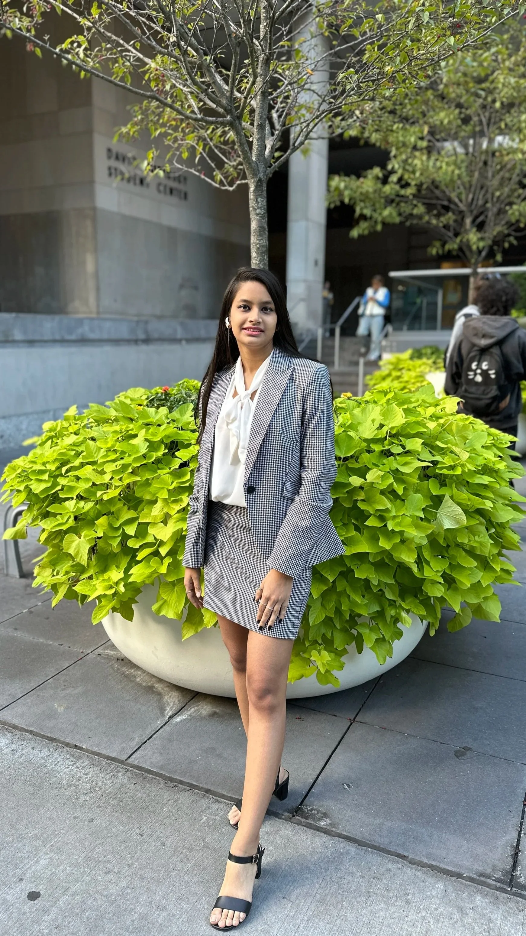 A woman in a checkered blazer and skirt standing in front of a large potted plant outside a building with the sign 'Davidson State Center'.