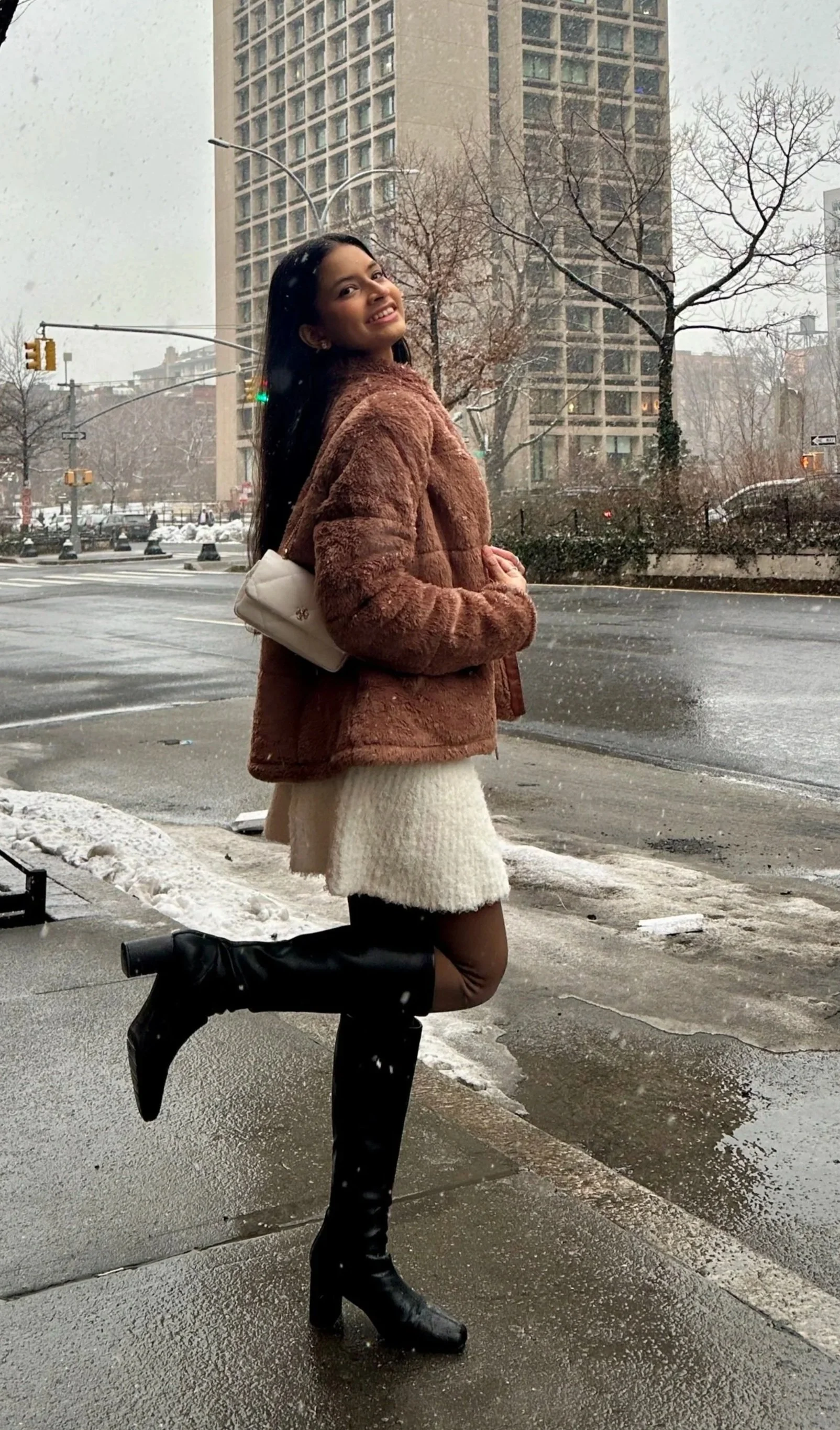 Young woman standing on city sidewalk during snowy weather, wearing a brown fur jacket, white skirt, and black knee-high boots, with buildings and bare trees in the background.