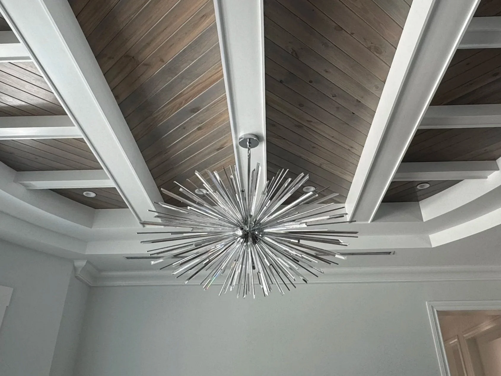 Modern chandelier with multiple metallic rods hanging from the ceiling of a room with a wooden-paneled ceiling and white walls.