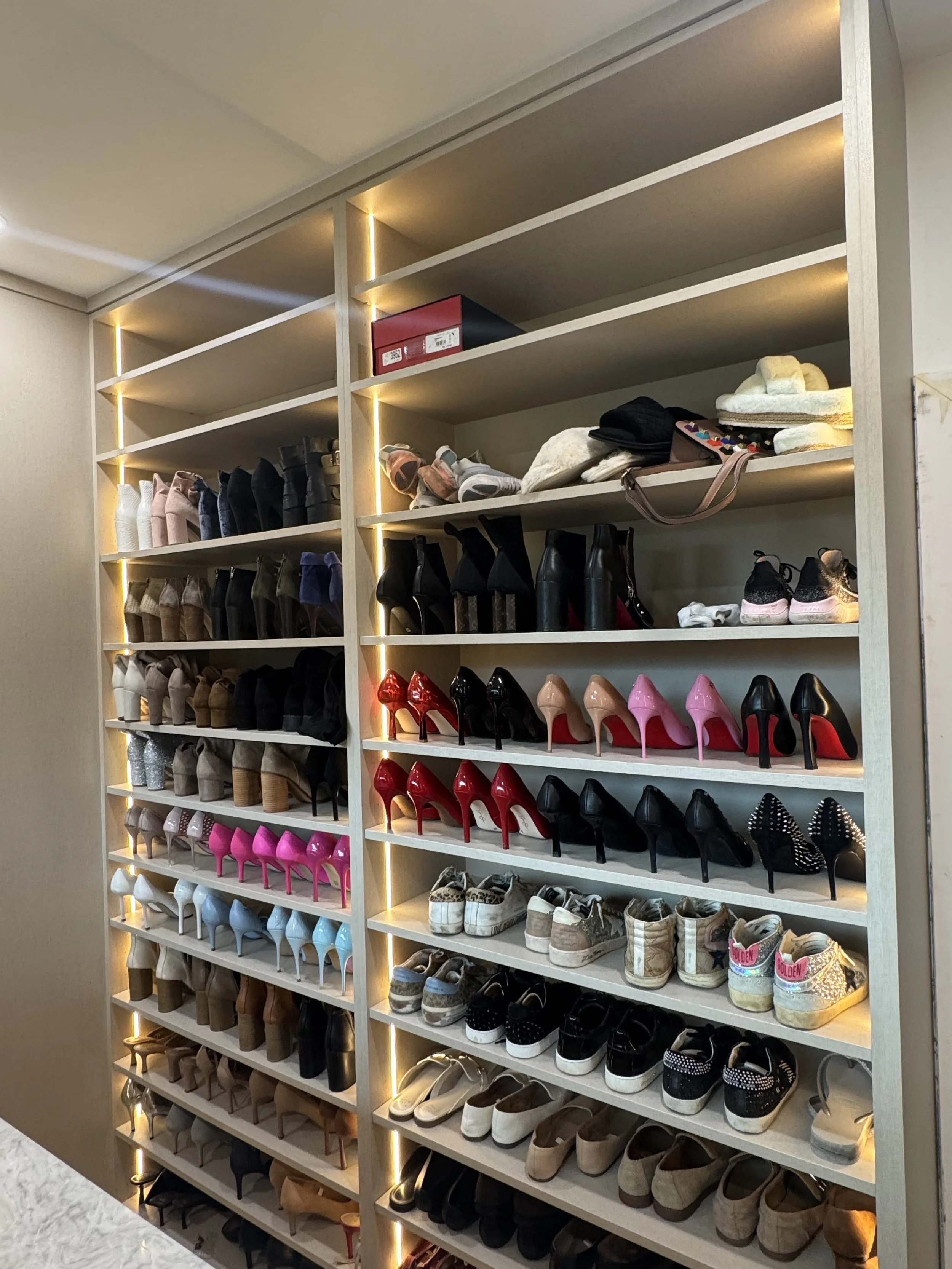 A wooden shoe rack with multiple shelves filled with various types of shoes, including high heels, sneakers, and boots, with LED strip lighting along the edges of the shelves.