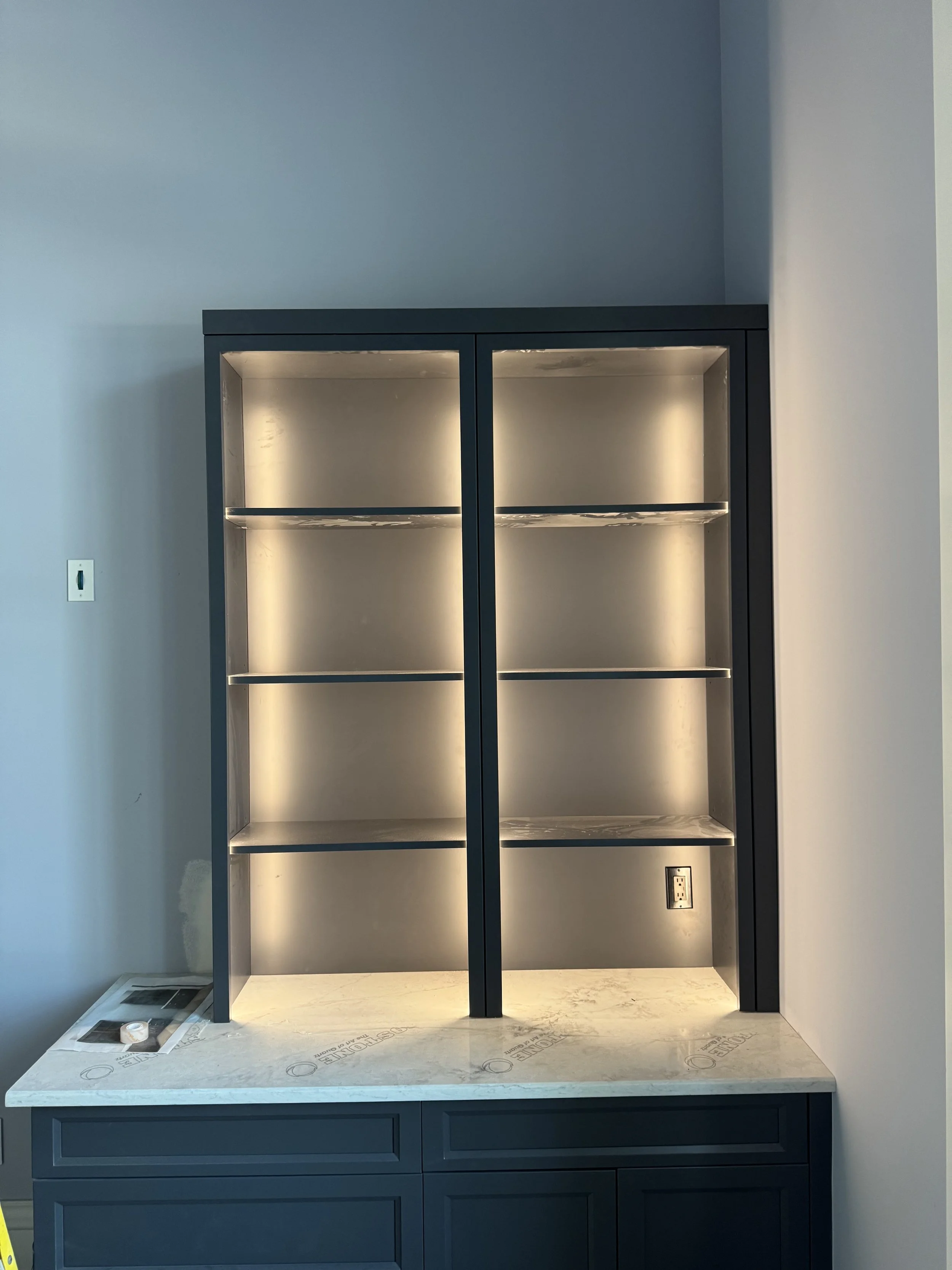 Empty built-in shelves with lighting, part of a cabinet with a marble countertop, in a room with light-colored walls and a power outlet.