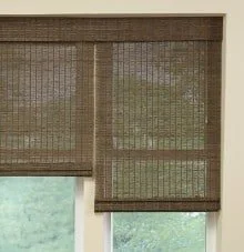 Close-up of brown woven window shades partially rolled up, revealing a view of trees and sky outside.