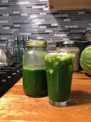 Two glasses of green juice or smoothie on a wooden kitchen countertop, with a jar and a watermelon in the background.