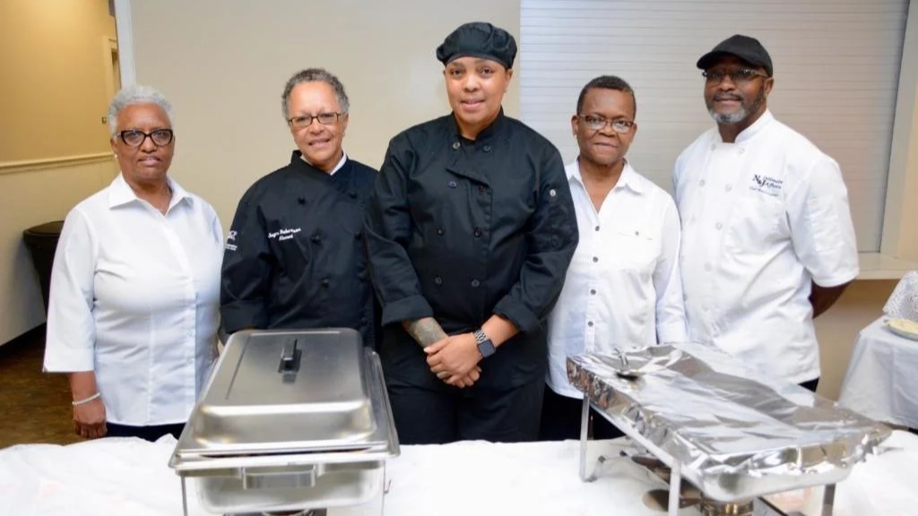 Chef Roxx and her team members are posing behind a table with serving trays at an event