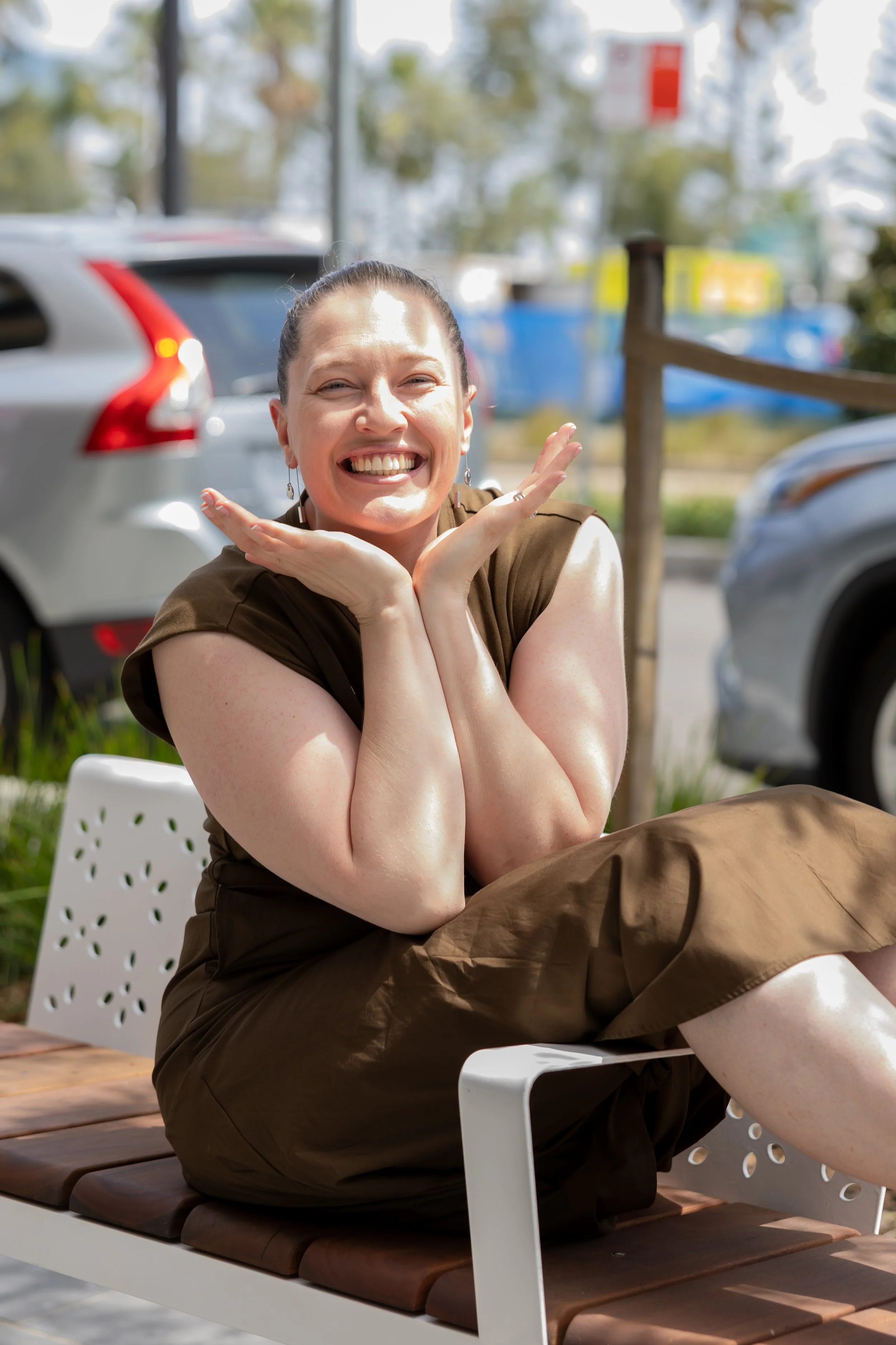 Smiling woman with short dark hair, wearing a brown dress, sitting on a white bench outdoors near parked cars, with trees and a clear sky in the background.