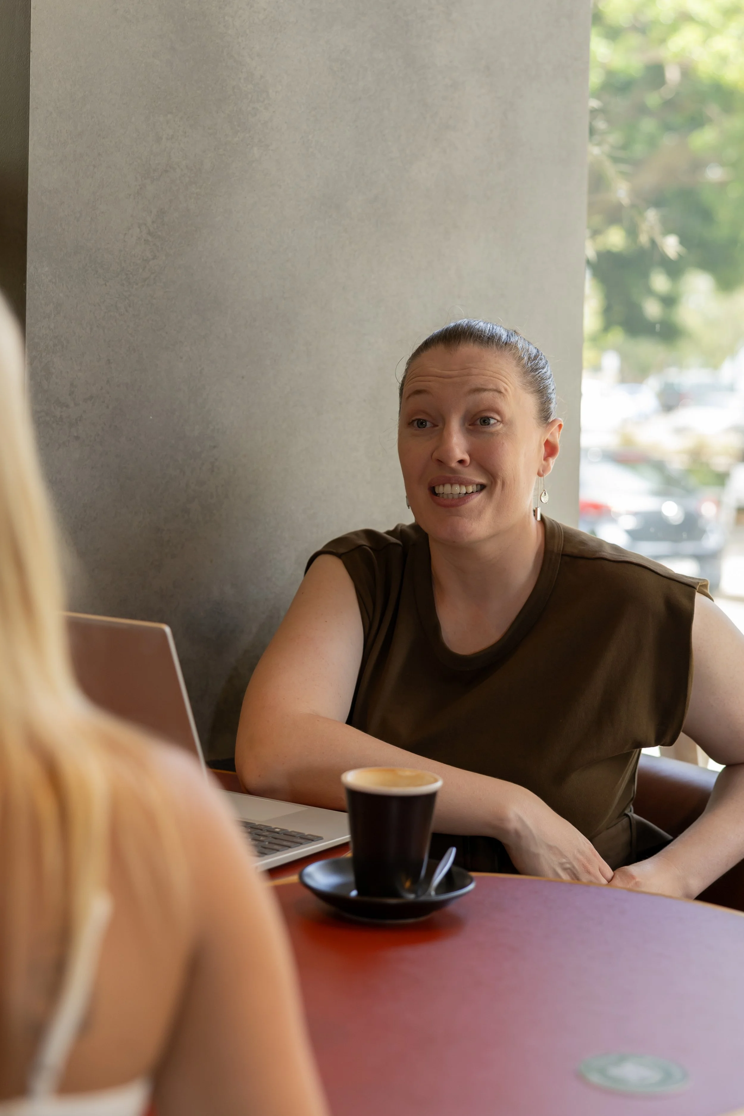 Teagan Pratt sitting at a table in a coffee shop, engaged in conversation, with a laptop and a cup of coffee in front of her.