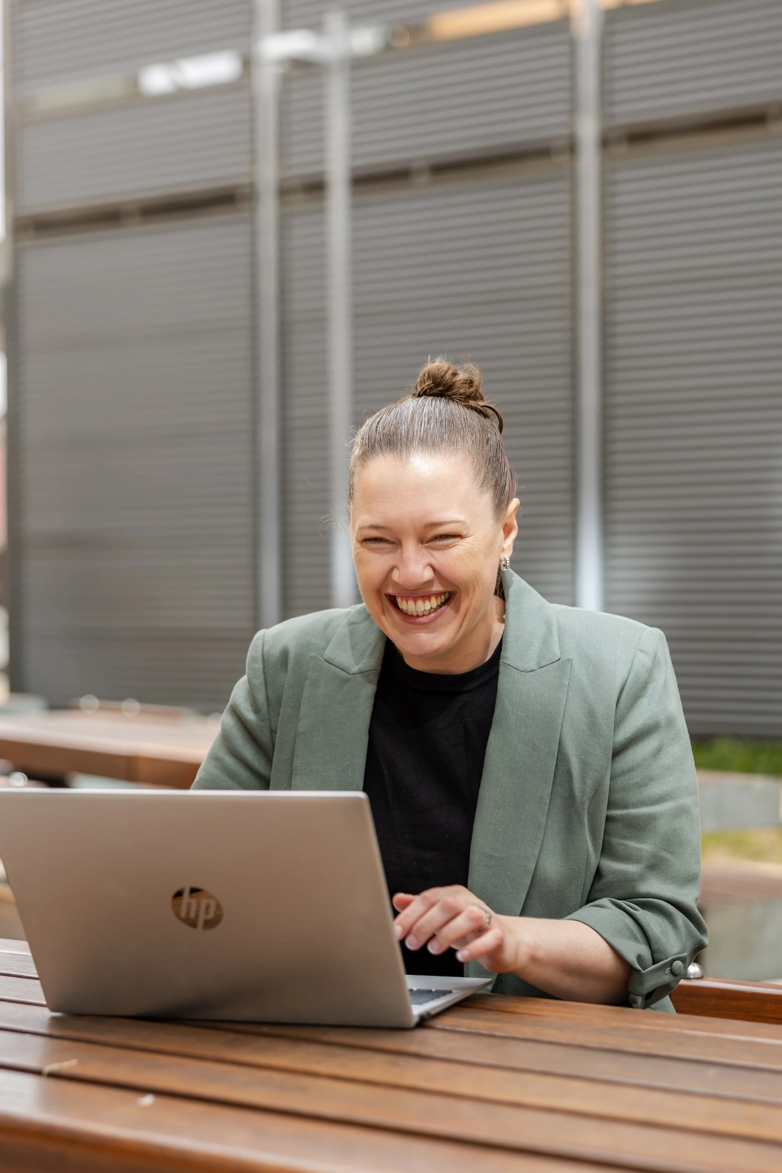 A woman with a bun hairstyle, wearing a green blazer, is sitting at a wooden outdoor table, smiling and laughing while looking at her laptop.