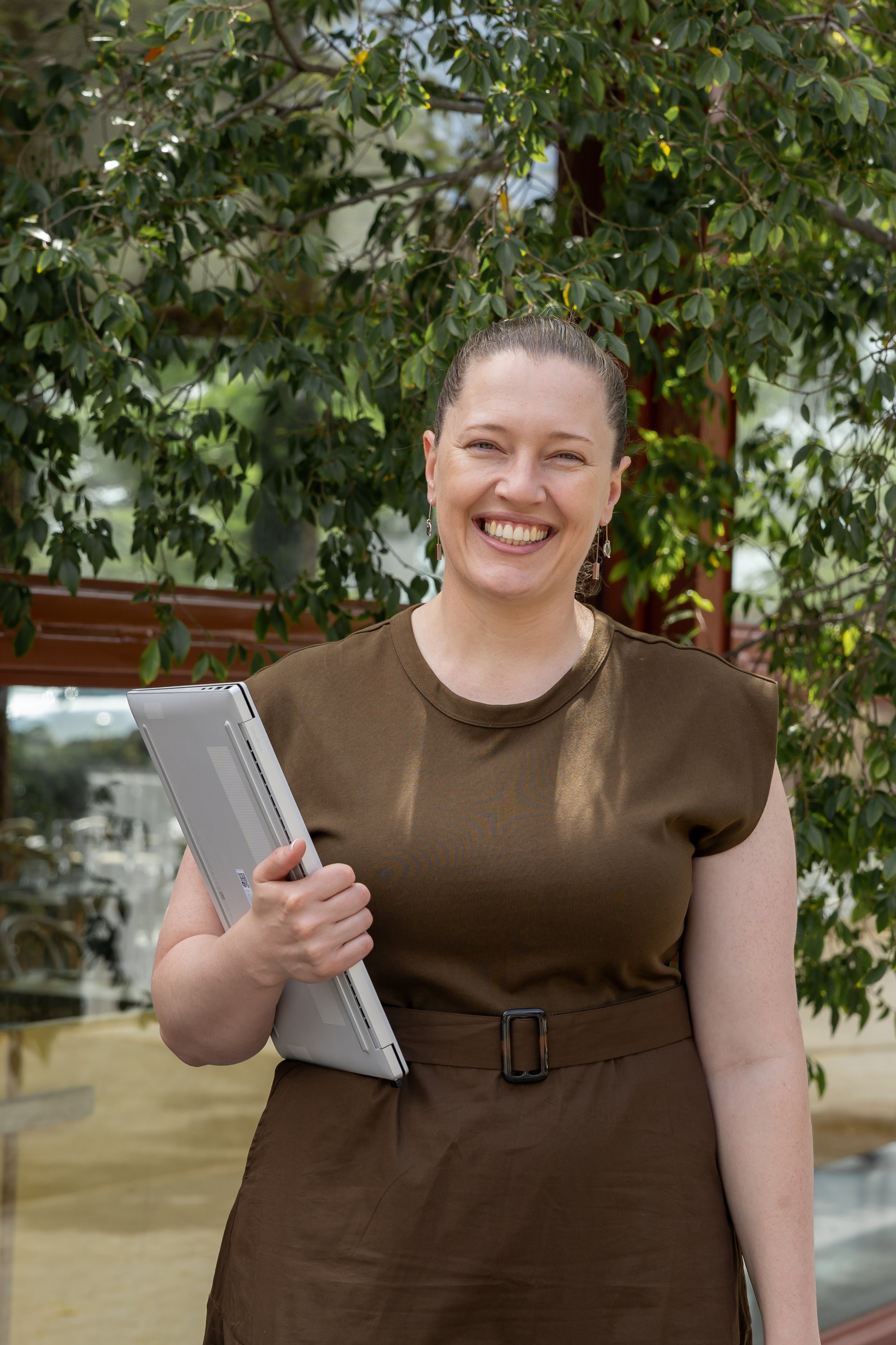 A woman smiling outdoors holding a laptop, standing in front of a leafy tree.
