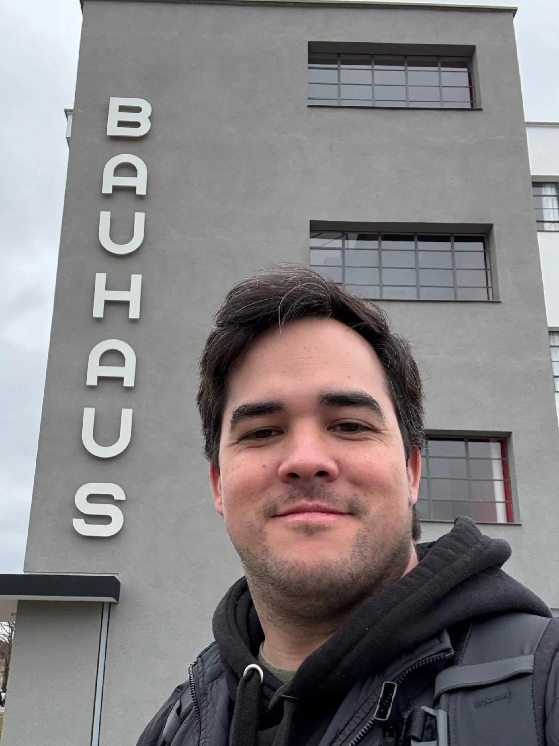 A man taking a selfie in front of a modern building with the word 'BAUHAUS' written vertically on the wall.