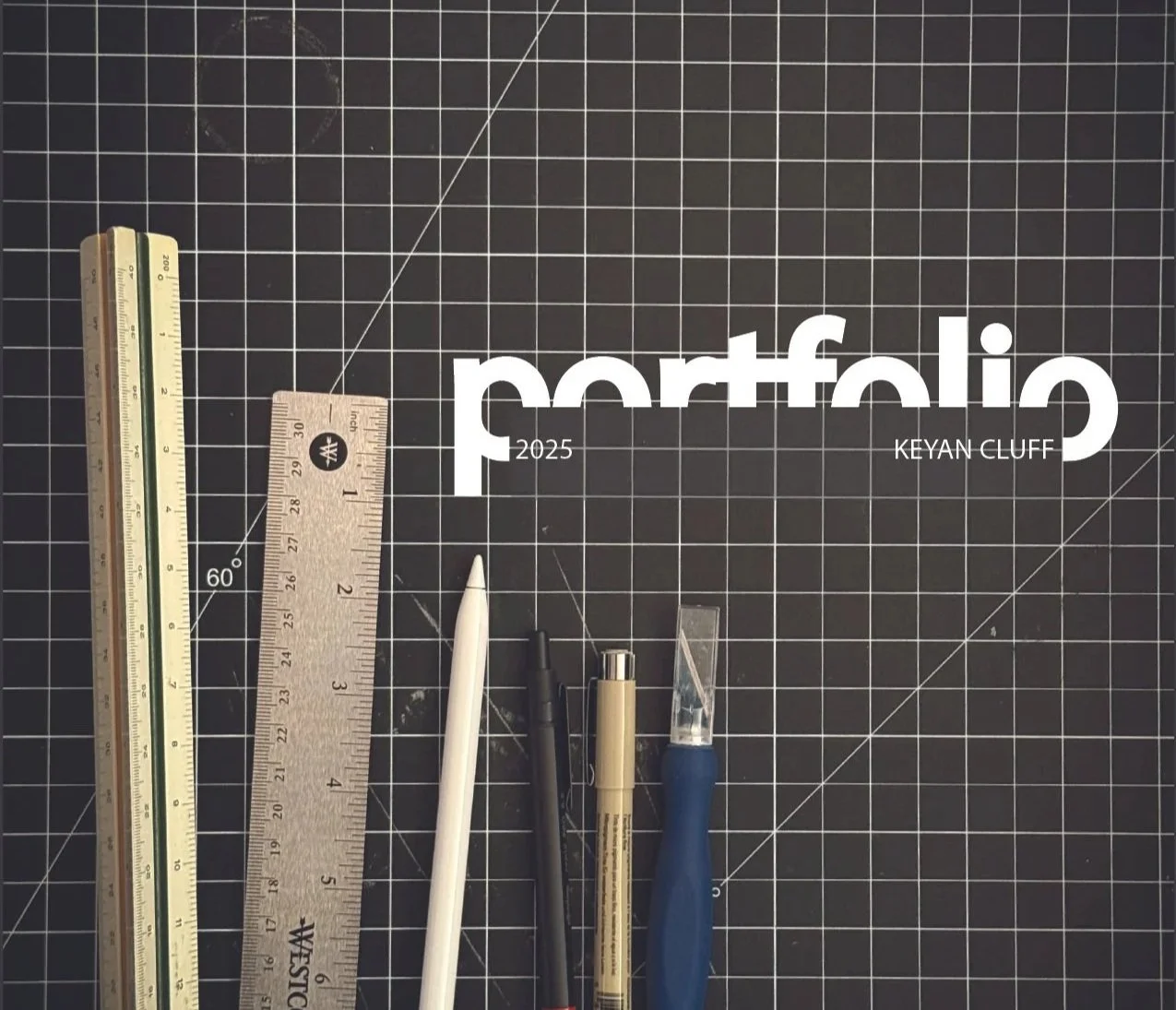 Multiple drawing tools including a yellow ruler, a clear plastic ruler, a white stylus, a black pen, a beige marker, and a blue craft knife on a black cutting mat with grid lines. The mat has white text that reads 'Portfolio 2025 Keyan Cluff'.
