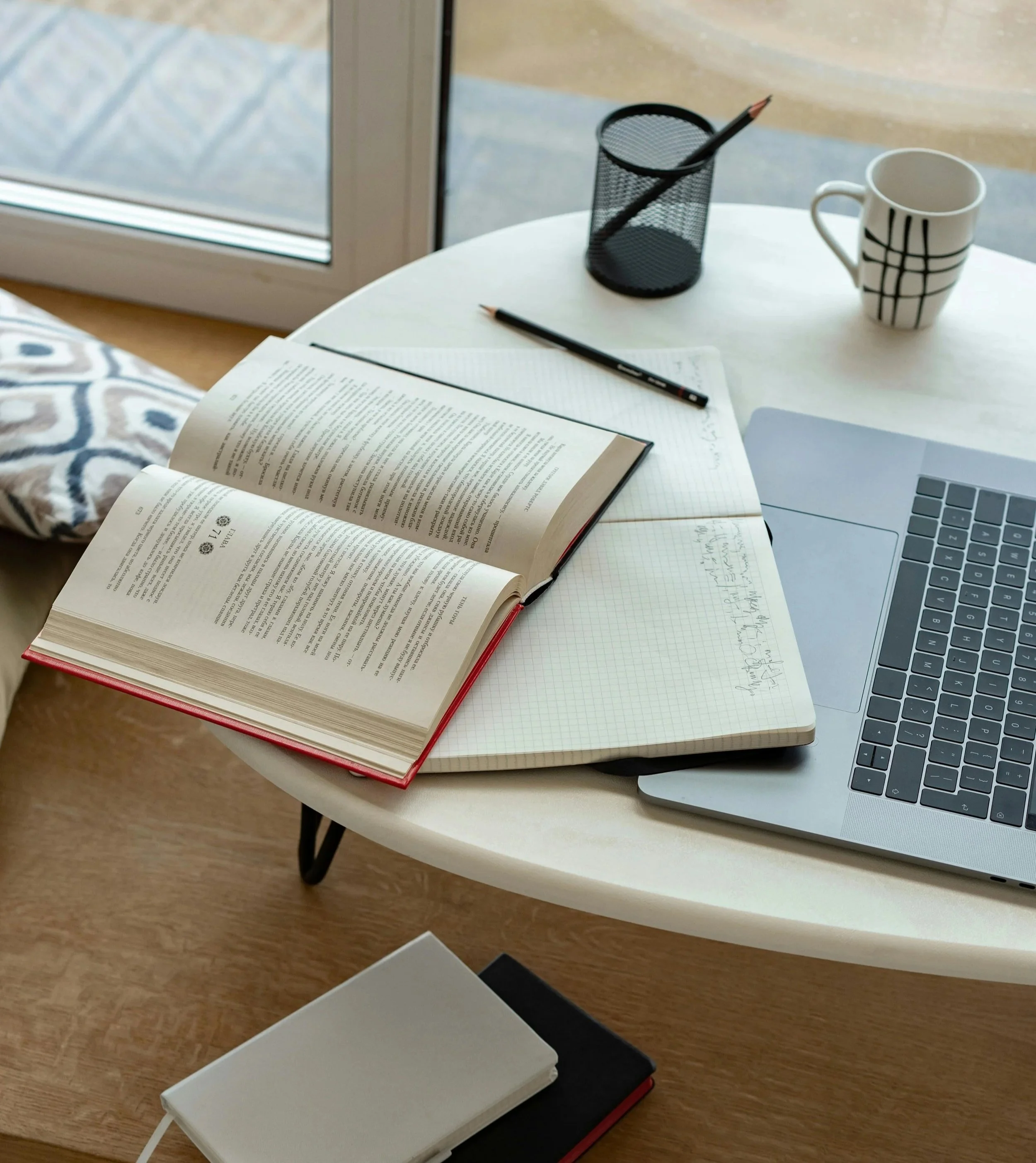 A round white coffee table with an open book, a closed notebook, a graph paper notebook, a laptop, a black pencil holder with a pencil, a mug with a black grid pattern, and a black pencil. The table is near a sliding glass door with a view of a balcony or patio and some objects outside.