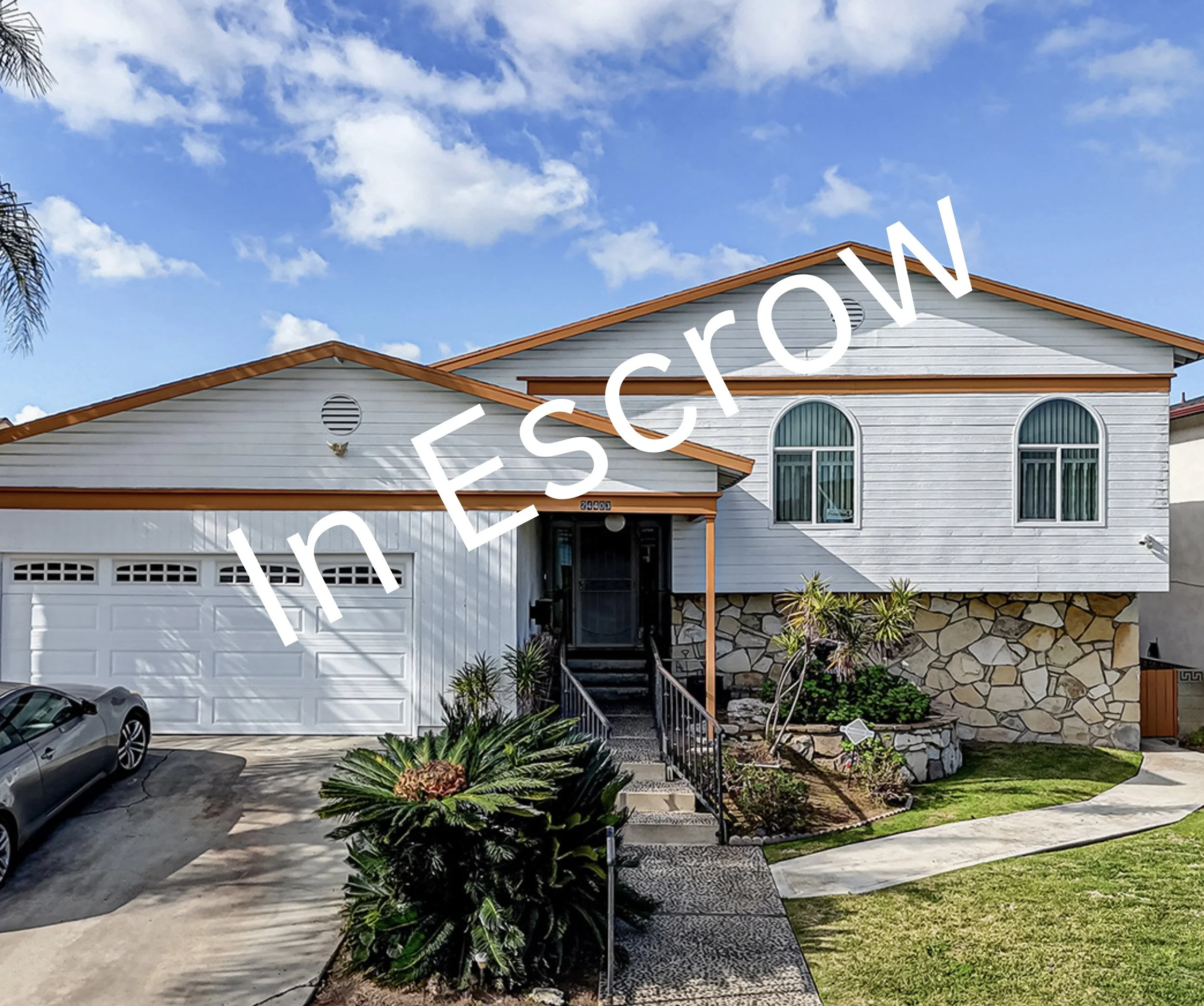 White house with a brown-tiled roof, attached garage, and front yard with plants and a driveway.