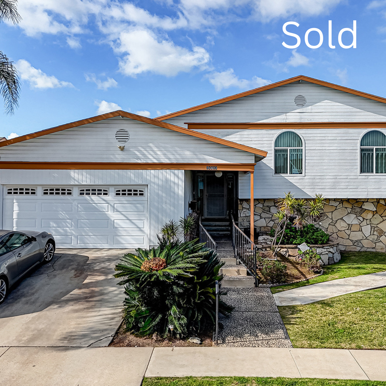 White house with a brown-tiled roof, attached garage, and front yard with plants and a driveway.