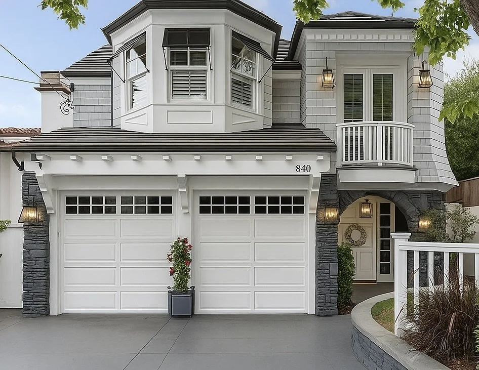 Front view of a modern two-story house with white siding, black roof, two garage doors, and front entrance under an archway, surrounded by greenery and outdoor lamps.