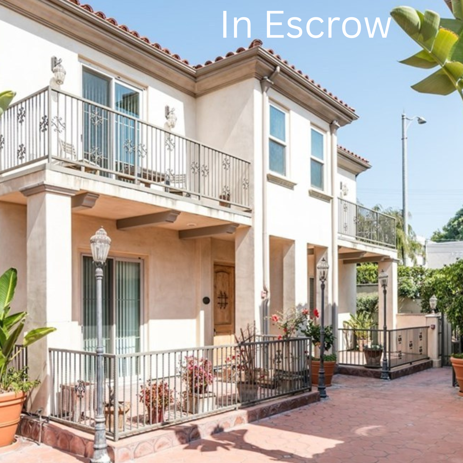 A two-story residential building with a light-colored stucco exterior and red tile roof, featuring a small balcony, potted plants, and a paved courtyard with decorative street lamps.