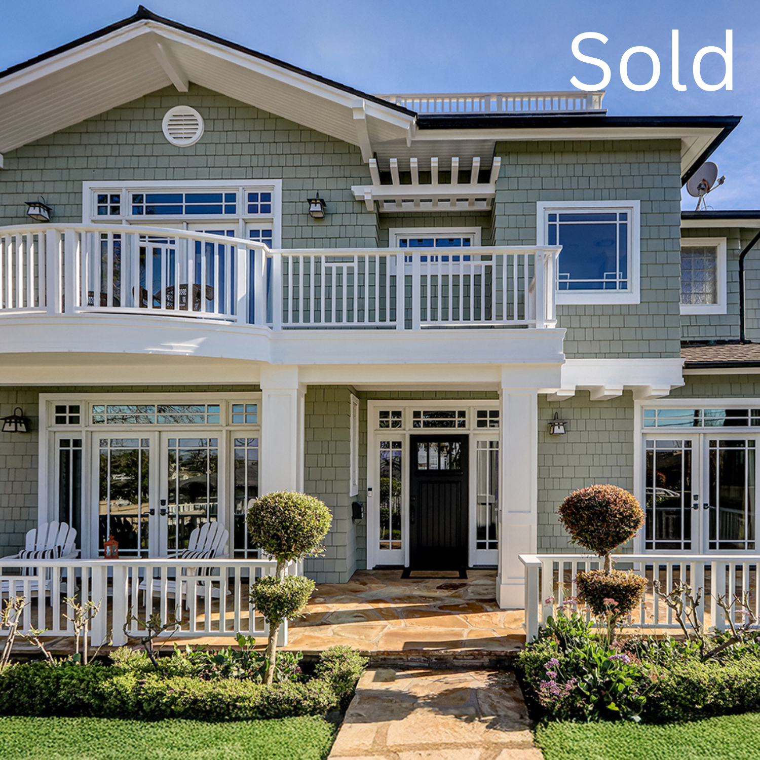 Front view of a multi-story house with a white balcony, gray siding, and a well-maintained garden with trimmed bushes and a stone walkway.