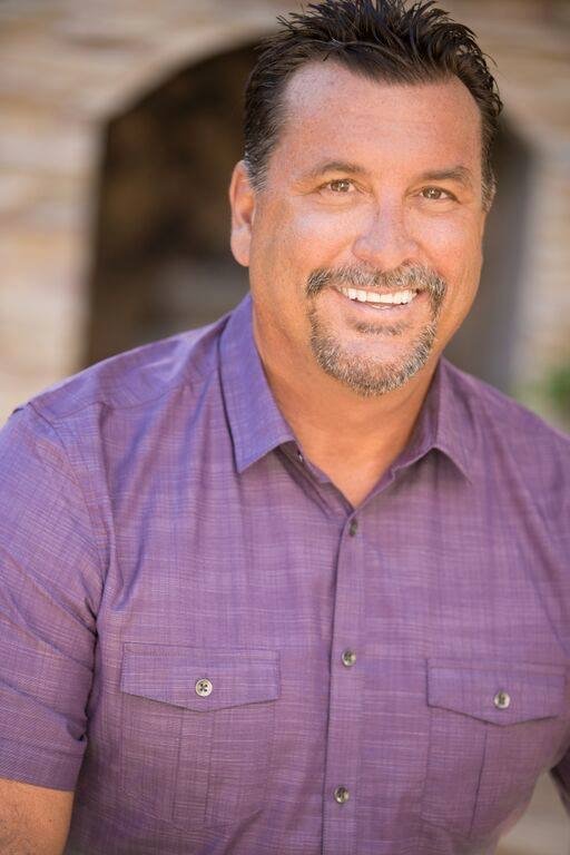 A smiling man with dark hair and a goatee, wearing a purple button-up shirt, outdoors with a stone wall in the background.