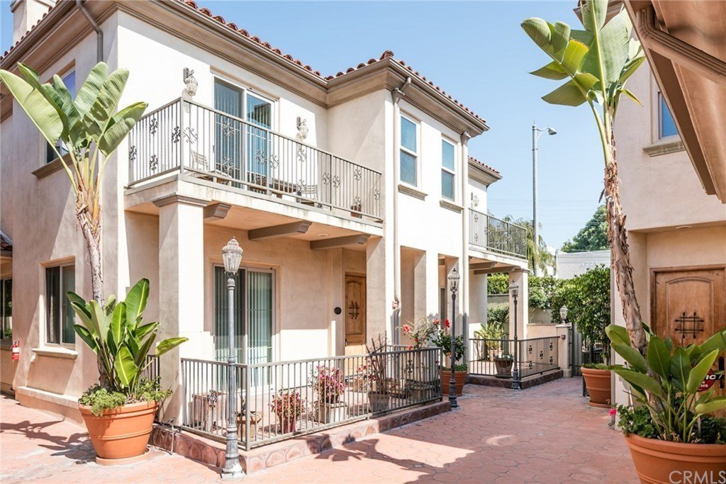 A two-story residential building with a light-colored stucco exterior and red tile roof, featuring a small balcony, potted plants, and a paved courtyard with decorative street lamps.