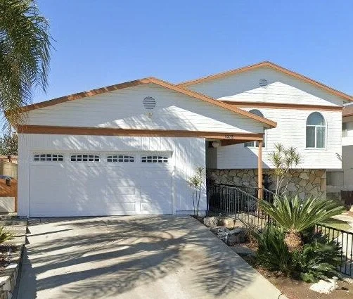 White house with a brown-tiled roof, attached garage, and front yard with plants and a driveway.