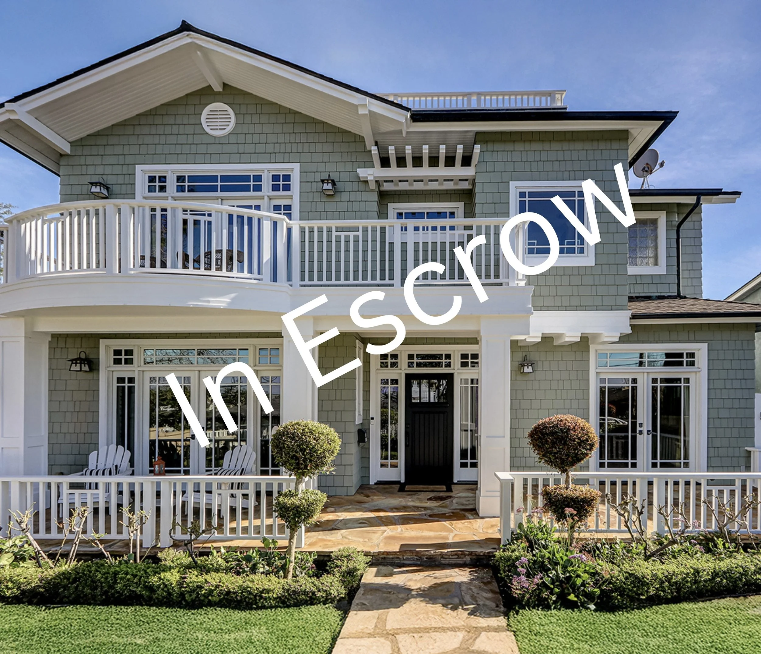 Front view of a multi-story house with a white balcony, gray siding, and a well-maintained garden with trimmed bushes and a stone walkway.
