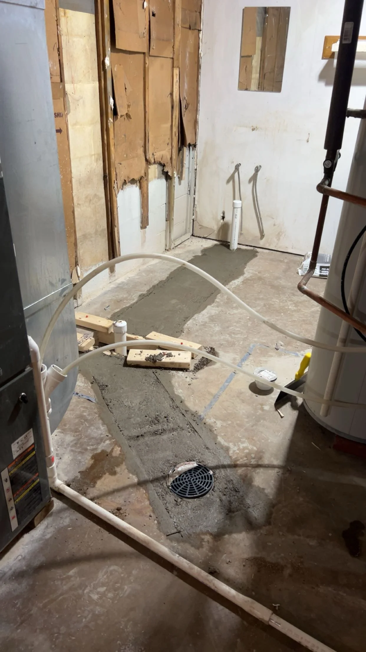 A bathroom under renovation showing exposed wooden wall studs, partially removed drywall, construction debris, and pipework. The floor is unfinished with visible concrete and a drain, with some gray cement patchwork.