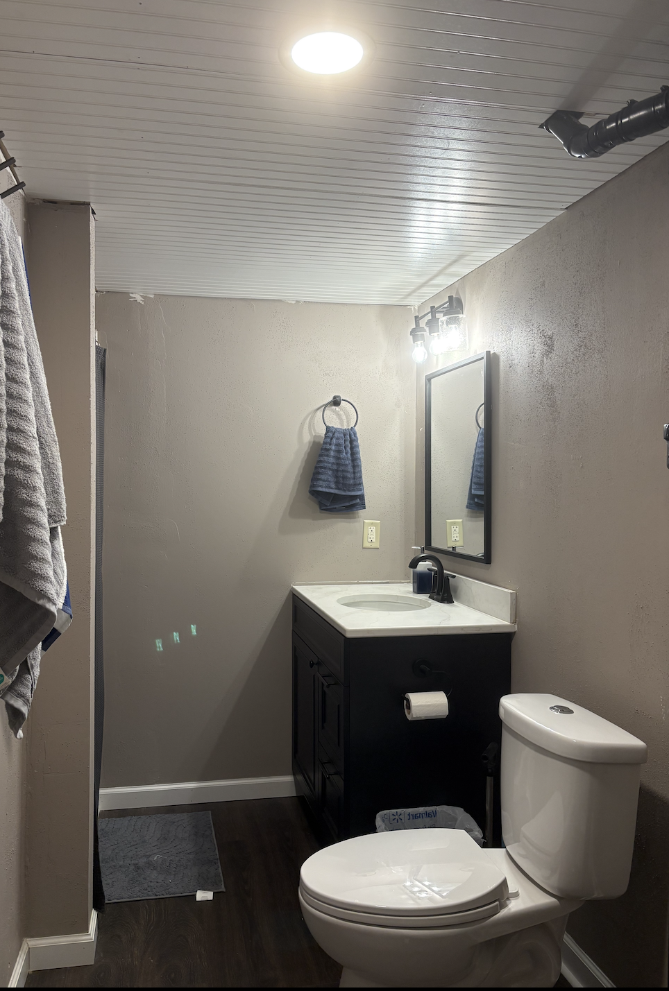A small bathroom with a white ceiling, beige walls, dark hardwood floor, a white toilet, and a black vanity with a white countertop and a sink. A mirror hangs above the vanity, with a wall-mounted light fixture above it. Towels hang on a ring on the 