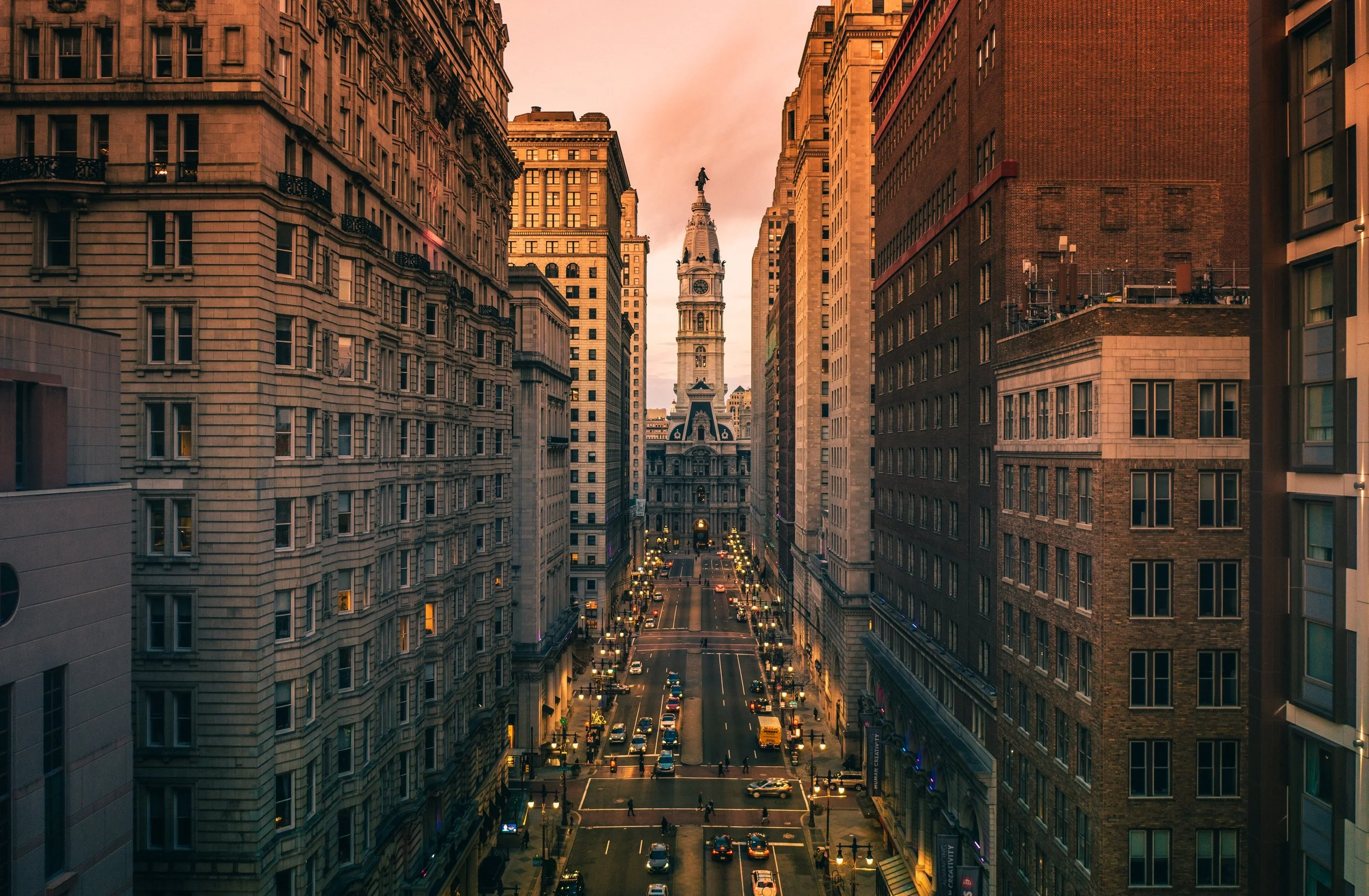 City street at dusk with tall historic buildings on both sides and a clock tower in the distance, with cars and pedestrians visible.