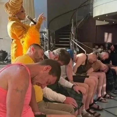 Six people sitting in chairs, leaning forward with heads down; behind them, a person in a bright orange and yellow costume holding a microphone.