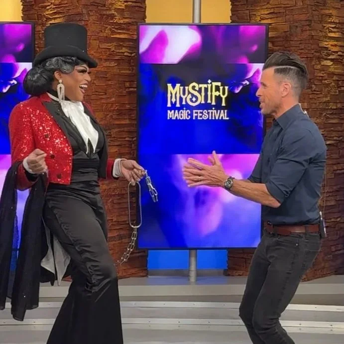 A woman dressed as a magician in a red sequined jacket, black top hat, and black pants, holding a chain, is performing a magic act on a man who is standing with his hands up in a playful manner. They are on a stage with a TV screen behind displaying 'Mystify Magic Festival.'