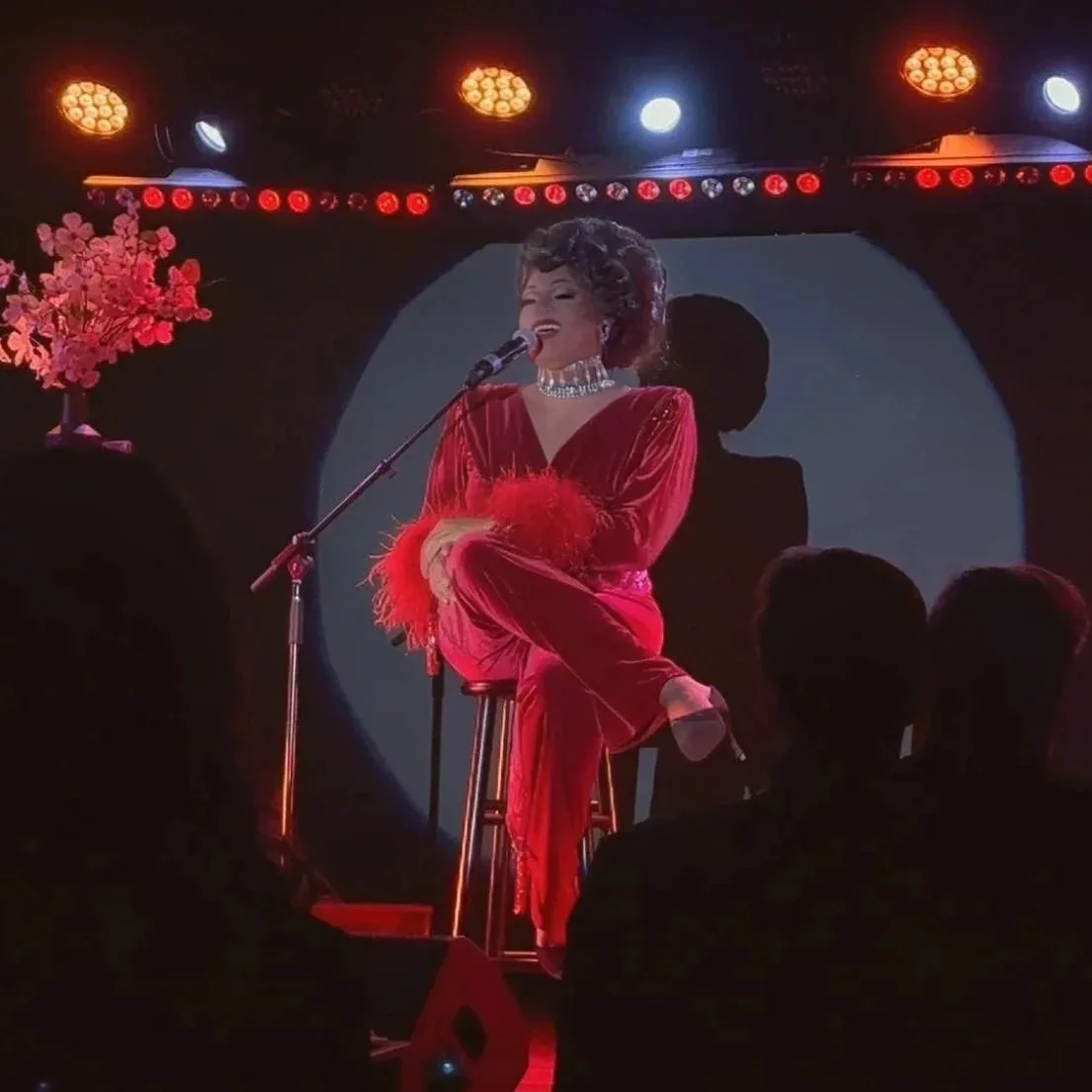 A female singer performing on stage, sitting on a stool, wearing a red outfit with feathered cuffs, and a sparkling choker, with stage lights and a silhouette backdrop.