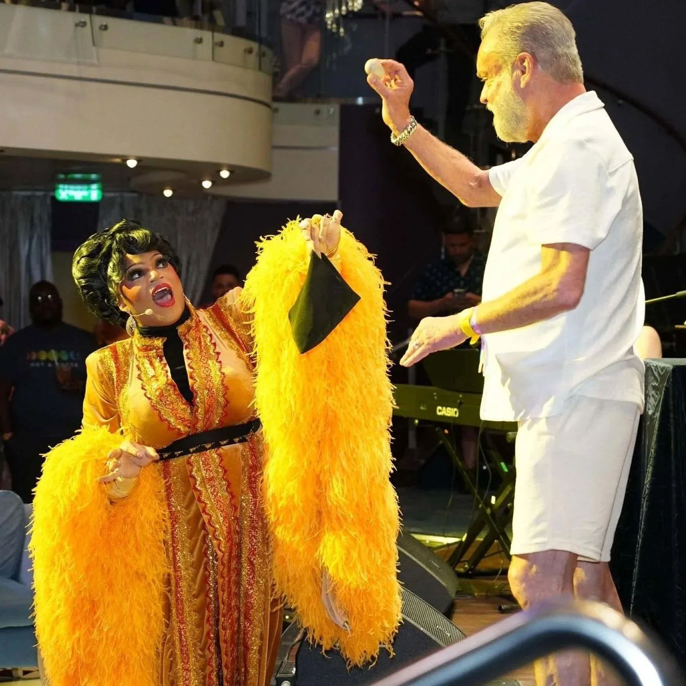 A performer in a vibrant yellow and orange costume with feathers, singing passionately on stage. An older man in a white outfit is holding a microphone, possibly engaging in a performance or interaction. The scene appears lively with a musical keyboard in the background and audience members watching.