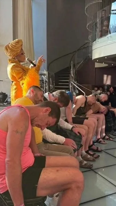A woman in a yellow outfit and wig, holding a microphone, stands on a stage with her arm raised. Several people are sitting in a row with their heads bowed, appearing to be asleep or resting.