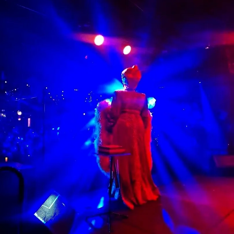 A performer wearing a long, glamorous gown and a hat, standing on stage under colorful blue and red lighting.