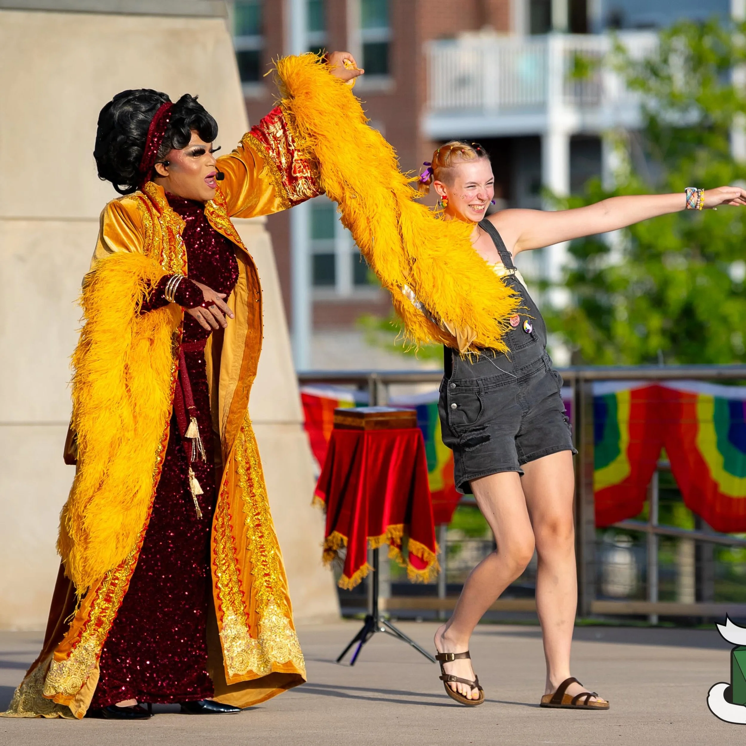 Performer dressed in a colorful, glittery costume with a yellow feather boa and a red sequined dress, dancing with a young girl on an outdoor stage, smiling and extending her arm, in front of a colorful tent and urban background.
