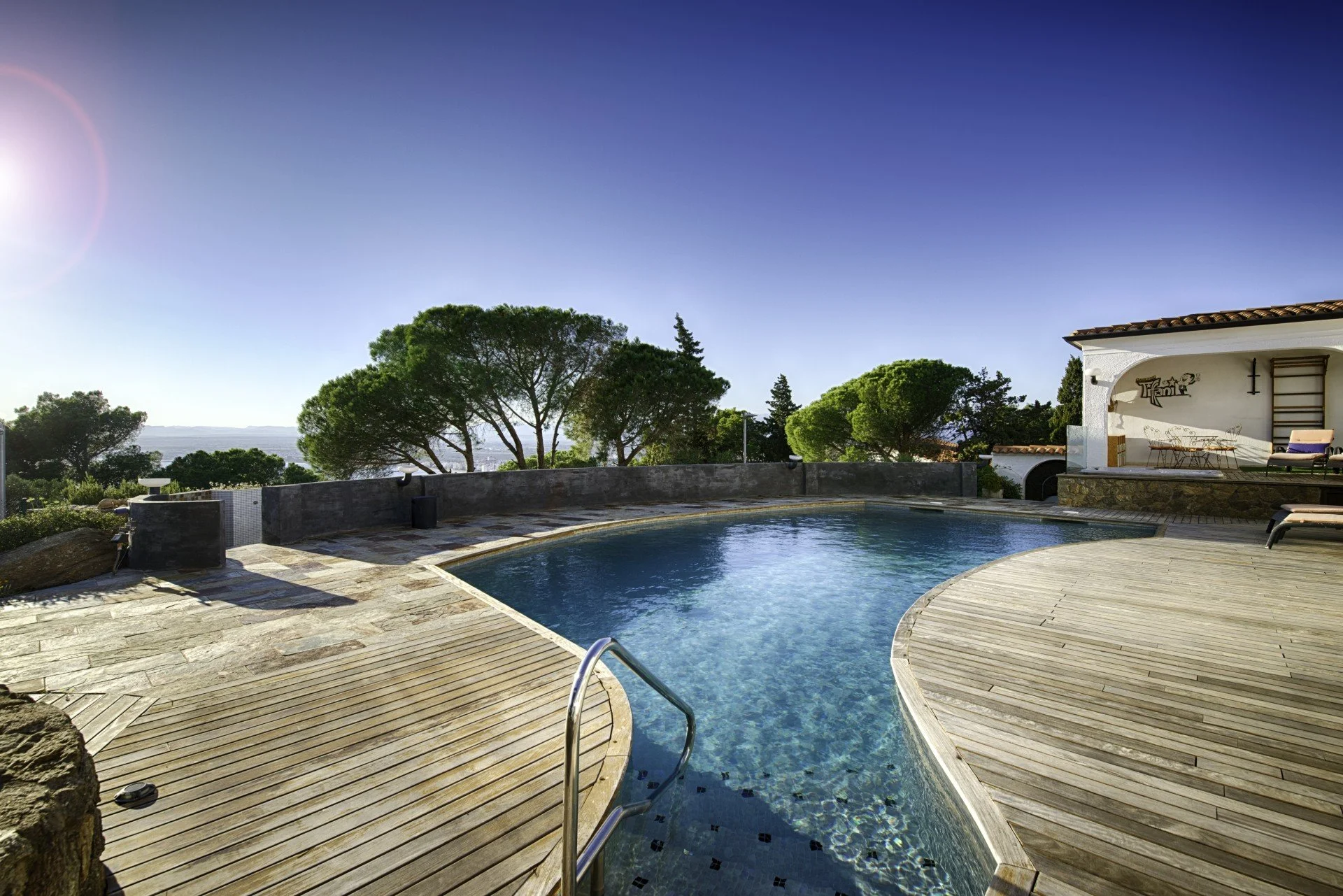 Poolside area with a curved swimming pool, surrounded by wooden decking, with trees and a white building with outdoor furniture in the background under a clear blue sky.