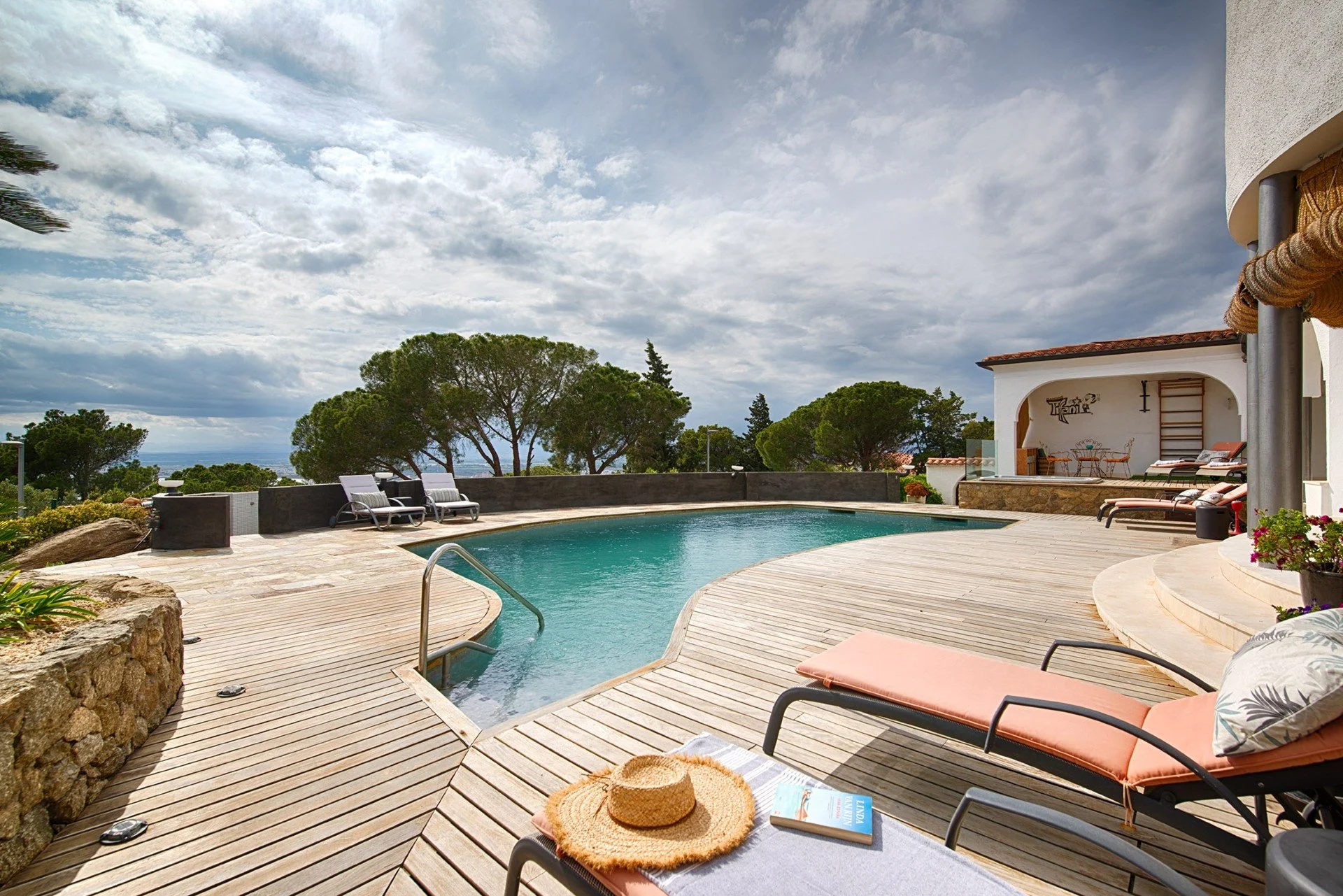 Poolside patio area with lounge chairs, a straw hat, a magazine, and a straw mat on a chair, overlooking a swimming pool surrounded by wooden decking and lush green trees under a partly cloudy sky.