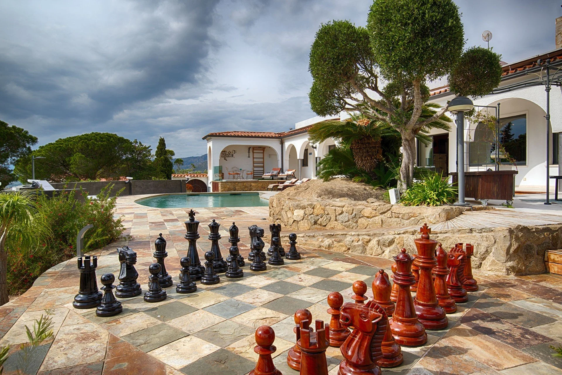 View of a luxurious backyard with a large chess set, a swimming pool, lush greenery, and white Mediterranean-style house with an outdoor patio under a cloudy sky.