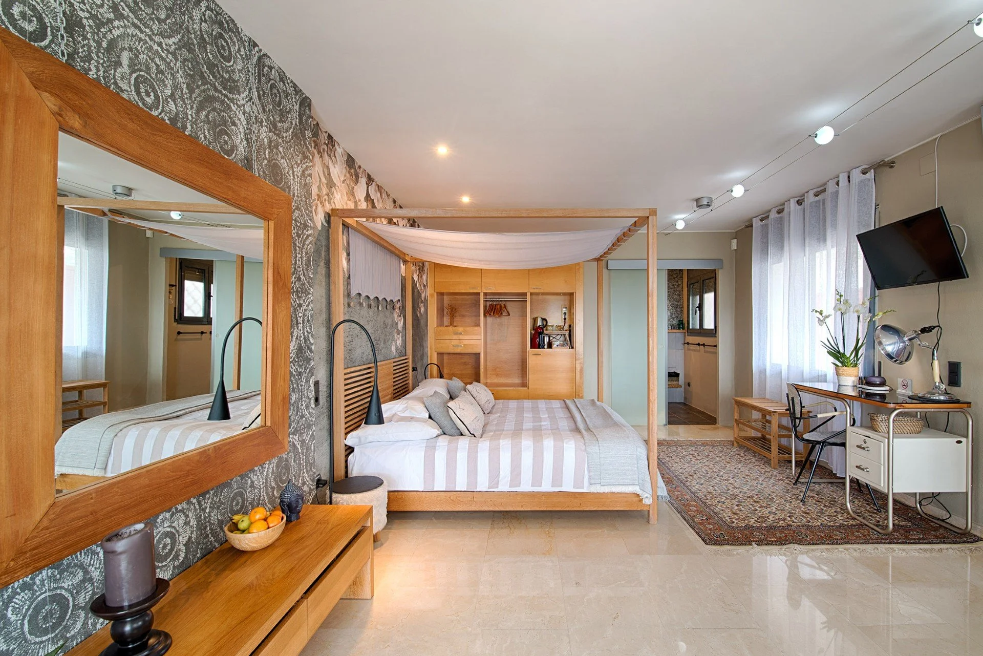 The Family Room at Casa de l'Albada, Roses. A modern hotel room with a bed, decorative wooden frame, patterned wallpaper, mirror, desk with a chair, television, and window with curtains, in a bright and well-lit space.