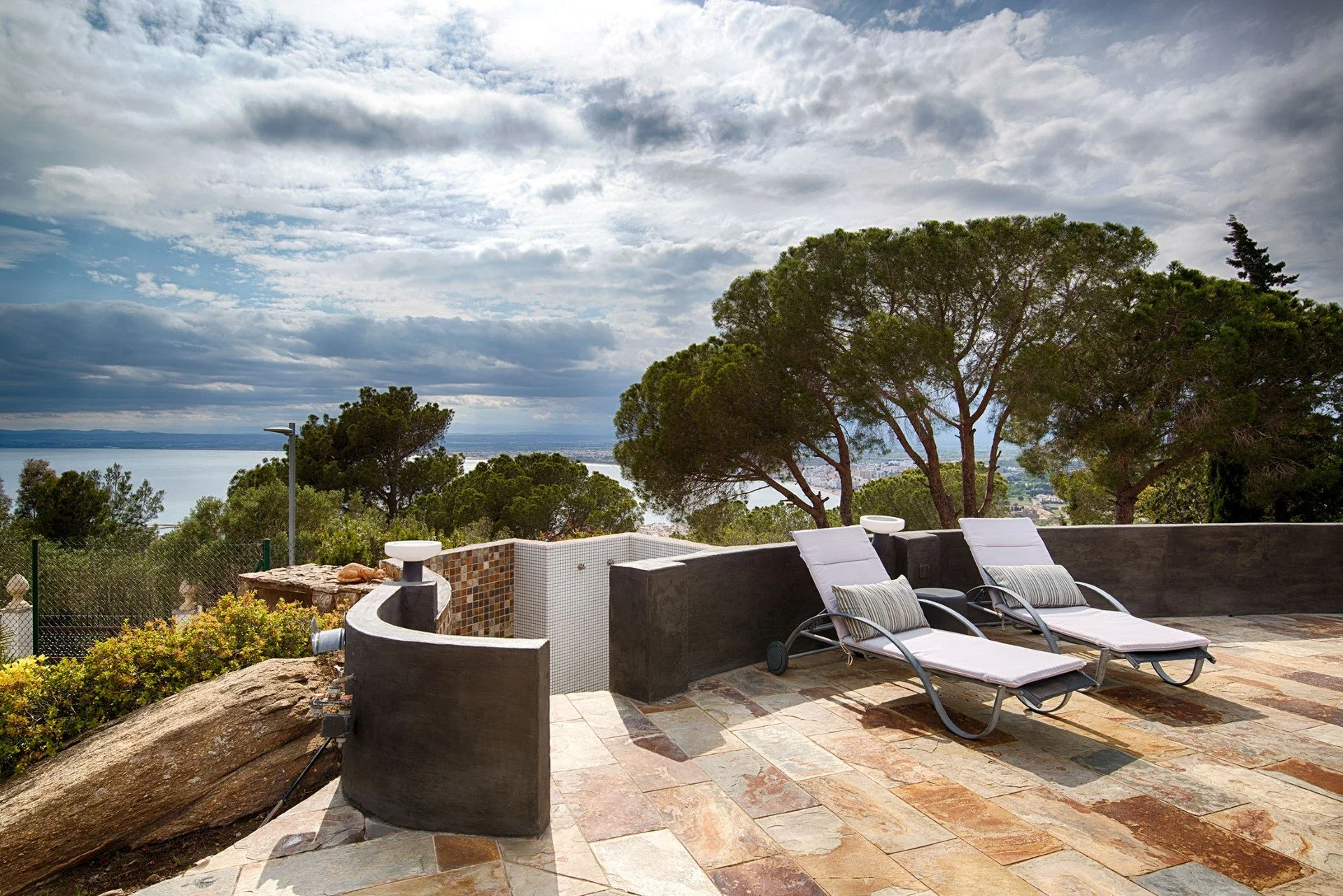 Outdoor patio with lounge chairs and a view of trees, water, and a cloudy sky.