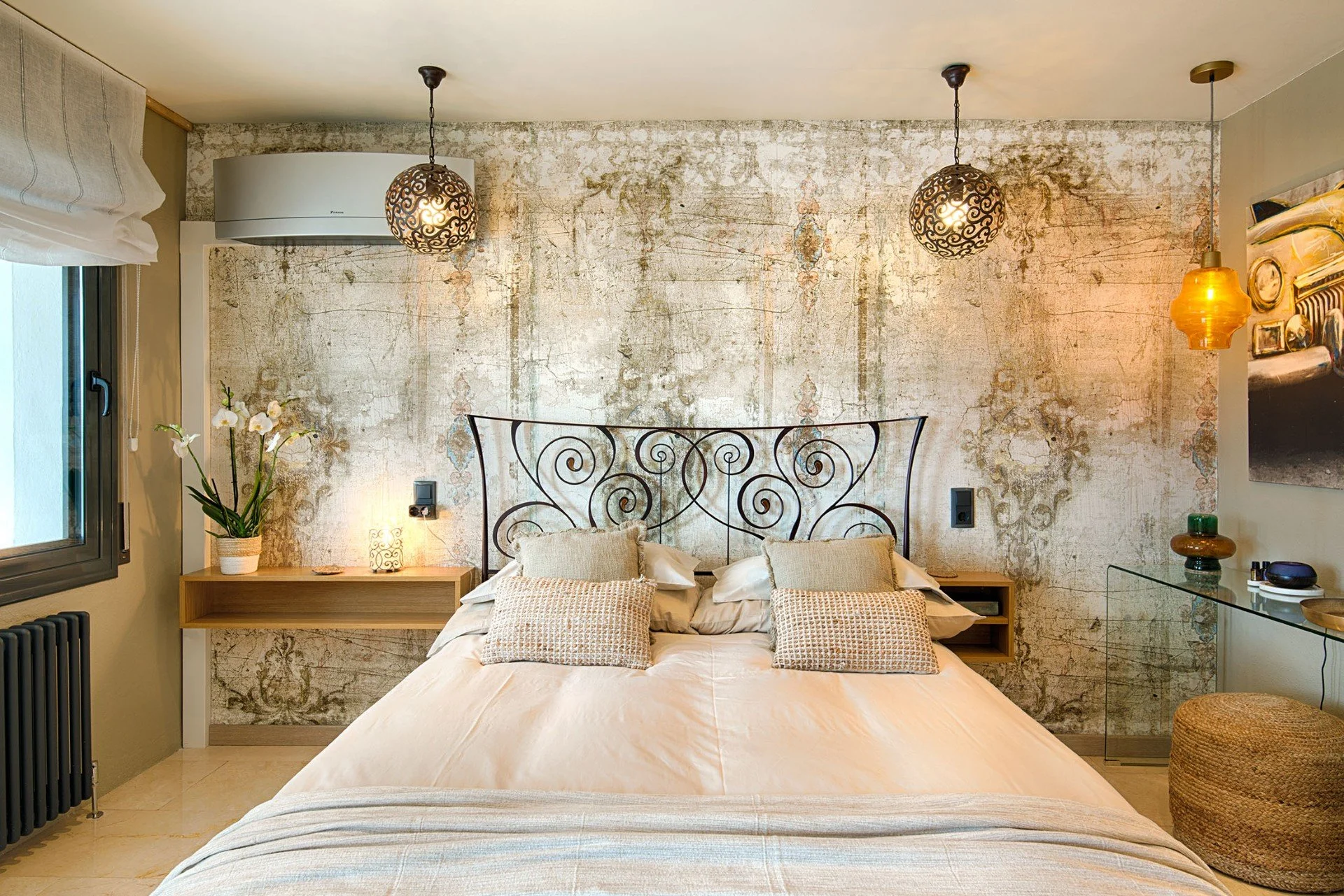 The Queen Room at Casa de l'Albada, Spain. Bedroom with a wrought iron bed, cozy pillows, a wooden nightstand with a potted plant, textured walls, hanging pendant lights, a window with a white curtain, and modern decor.