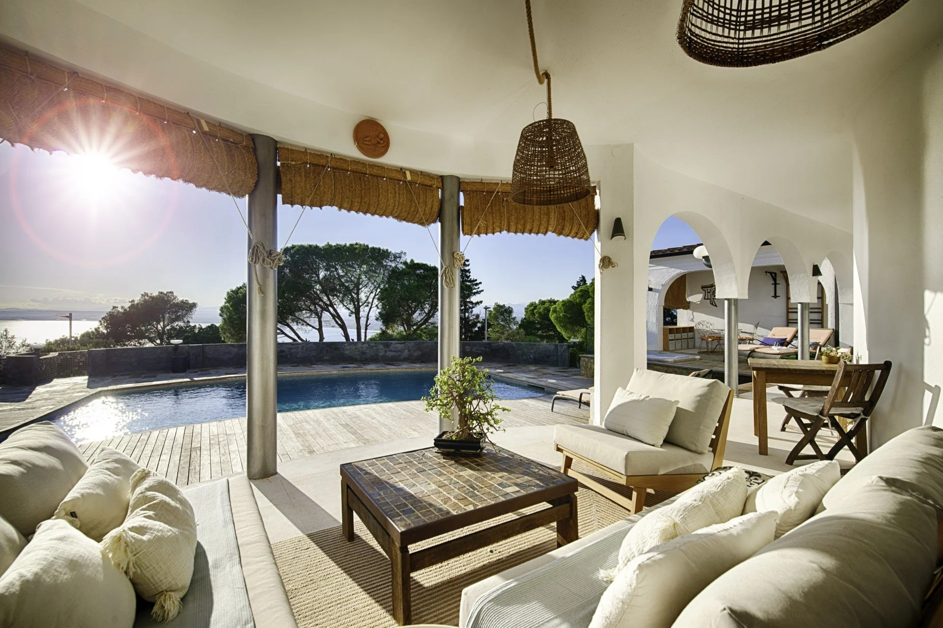 Outdoor patio with white cushioned sofas around a wooden coffee table, overlooking a swimming pool and trees under a clear sky, partially shaded by a thatched roof and wicker pendant lights.