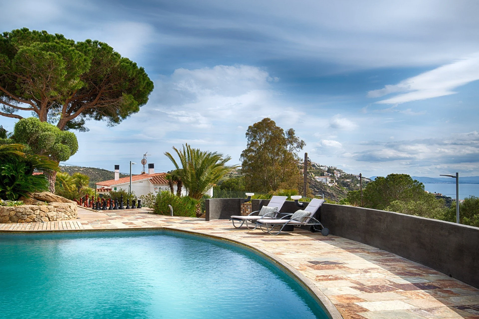 A backyard with a swimming pool, lounge chairs, trees, and a view of distant hills and water.