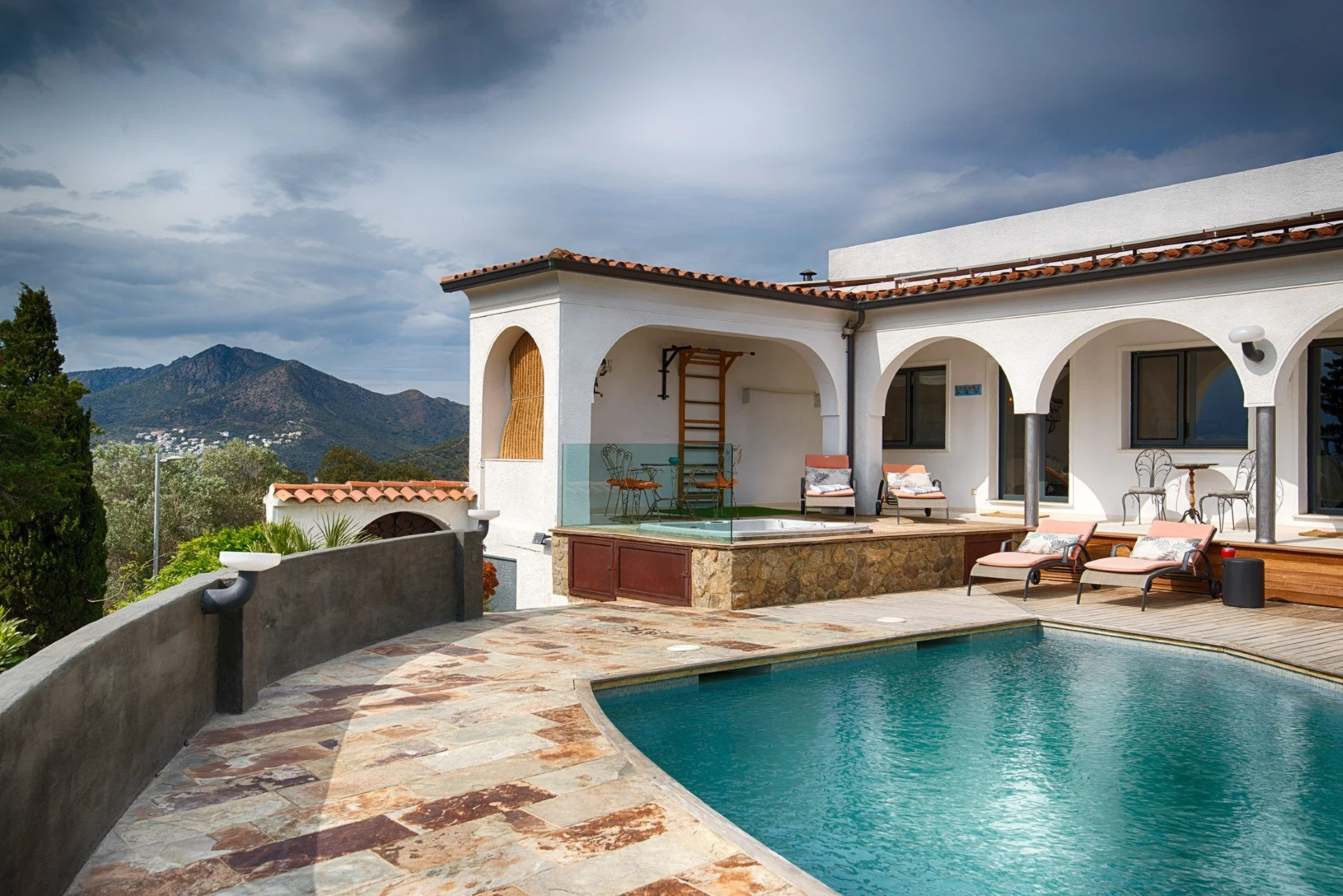 A backyard with a swimming pool, patio furniture, and scenic mountains in the background. White stucco house with arched windows and terracotta roof tiles.