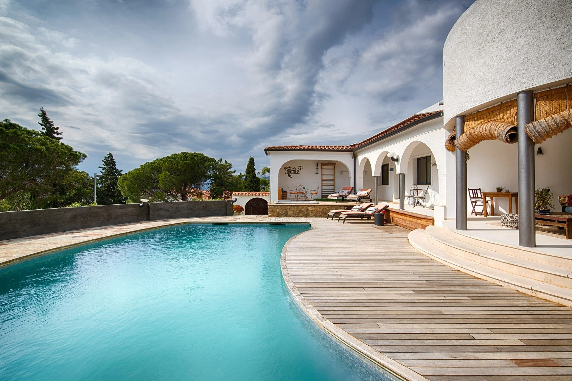A backyard with a swimming pool, surrounded by wooden decking and outdoor furniture, adjacent to a white house with arches and a red-tiled roof under a cloudy sky.