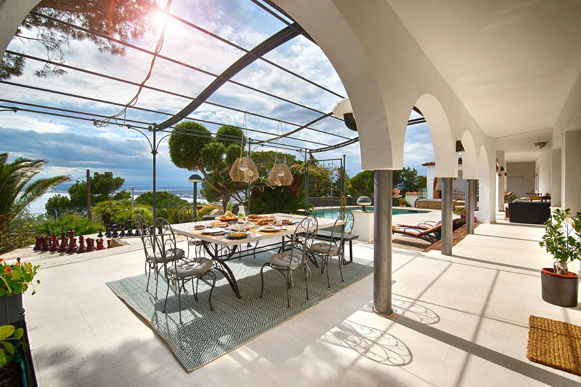 Outdoor patio with dining table set for breakfast, surrounded by greenery and overlooking a body of water, with a pool and lounge chairs in the background.