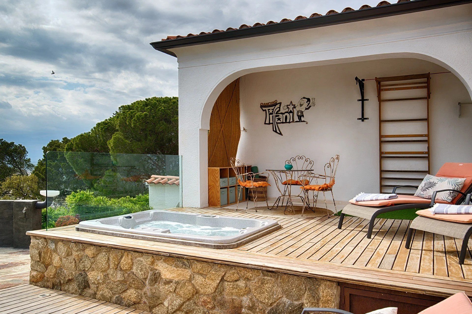 Outdoor patio with hot tub, lounge chairs with cushions, small dining table with four chairs, and a wall with decorative sign, under a cloudy sky.