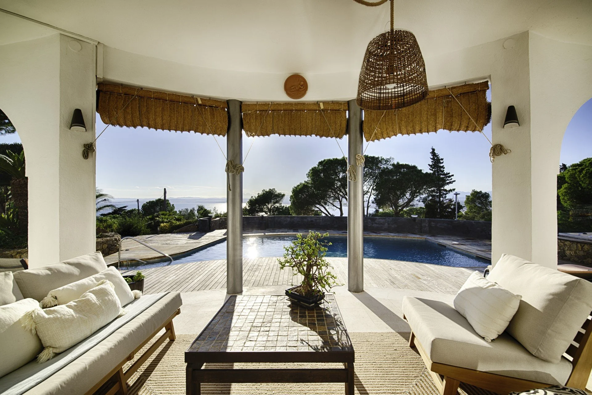 Outdoor patio area with white cushioned seating, a wooden coffee table, and a view of a swimming pool and trees in the background.